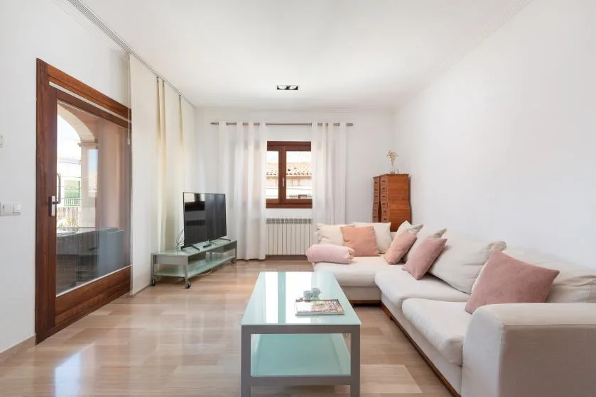 A bright living room with a pale sectional sofa, glass coffee table, and television.