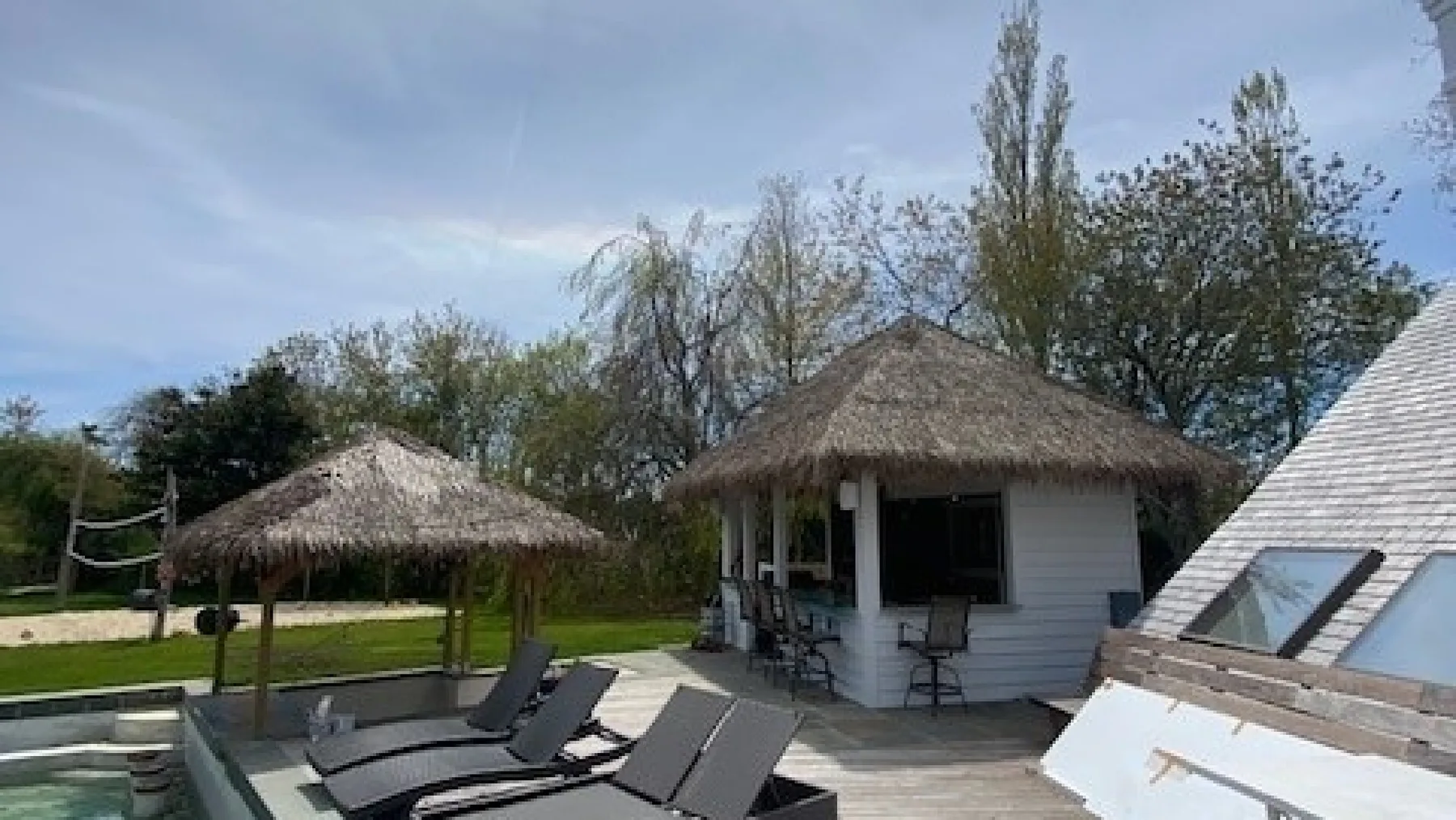 An outdoor patio with a thatched-roof gazebo, lounge seating, and a small white building.