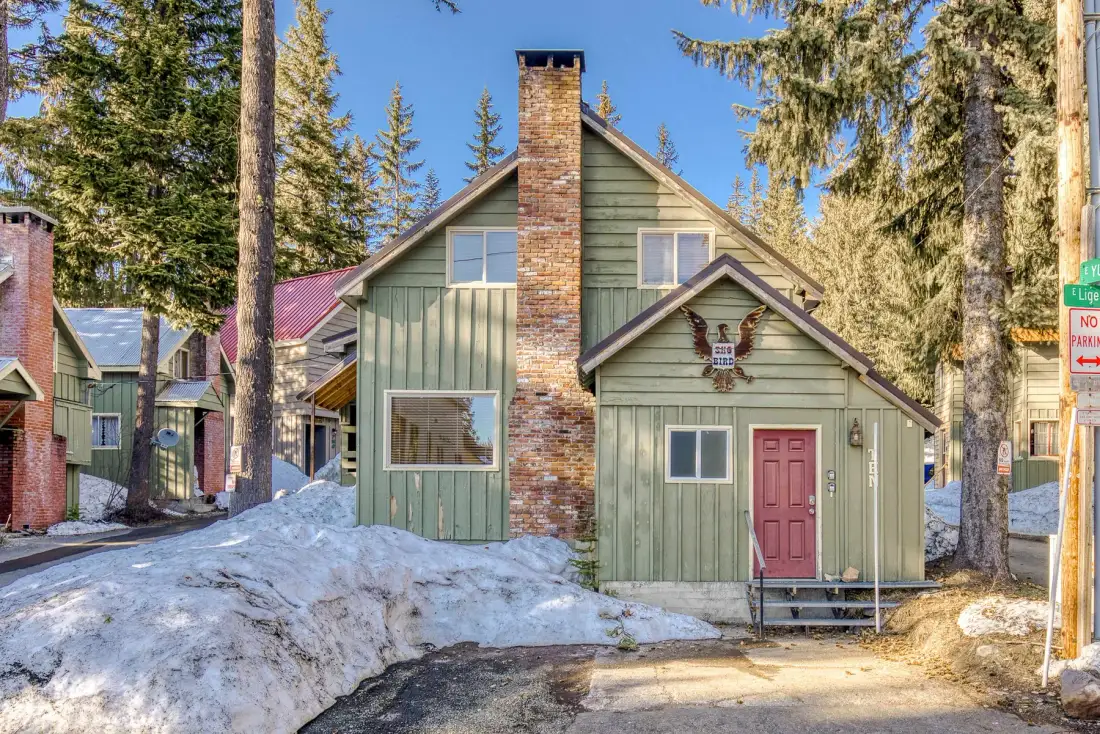 A green cabin with a brick chimney stands beside a snow-covered road.