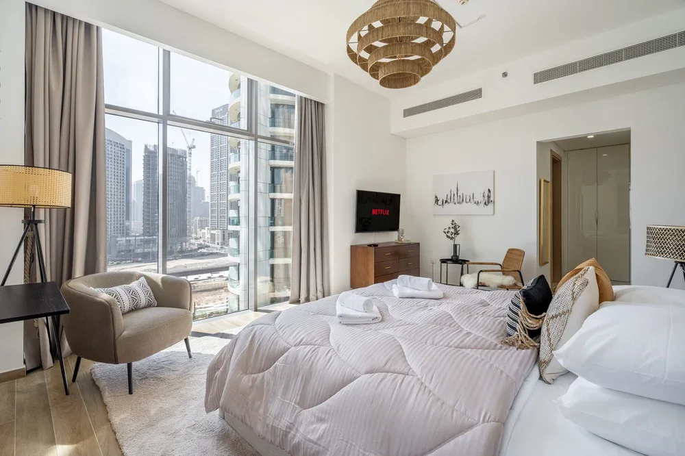 A bright bedroom with large windows, white bedding, and a seating area.
