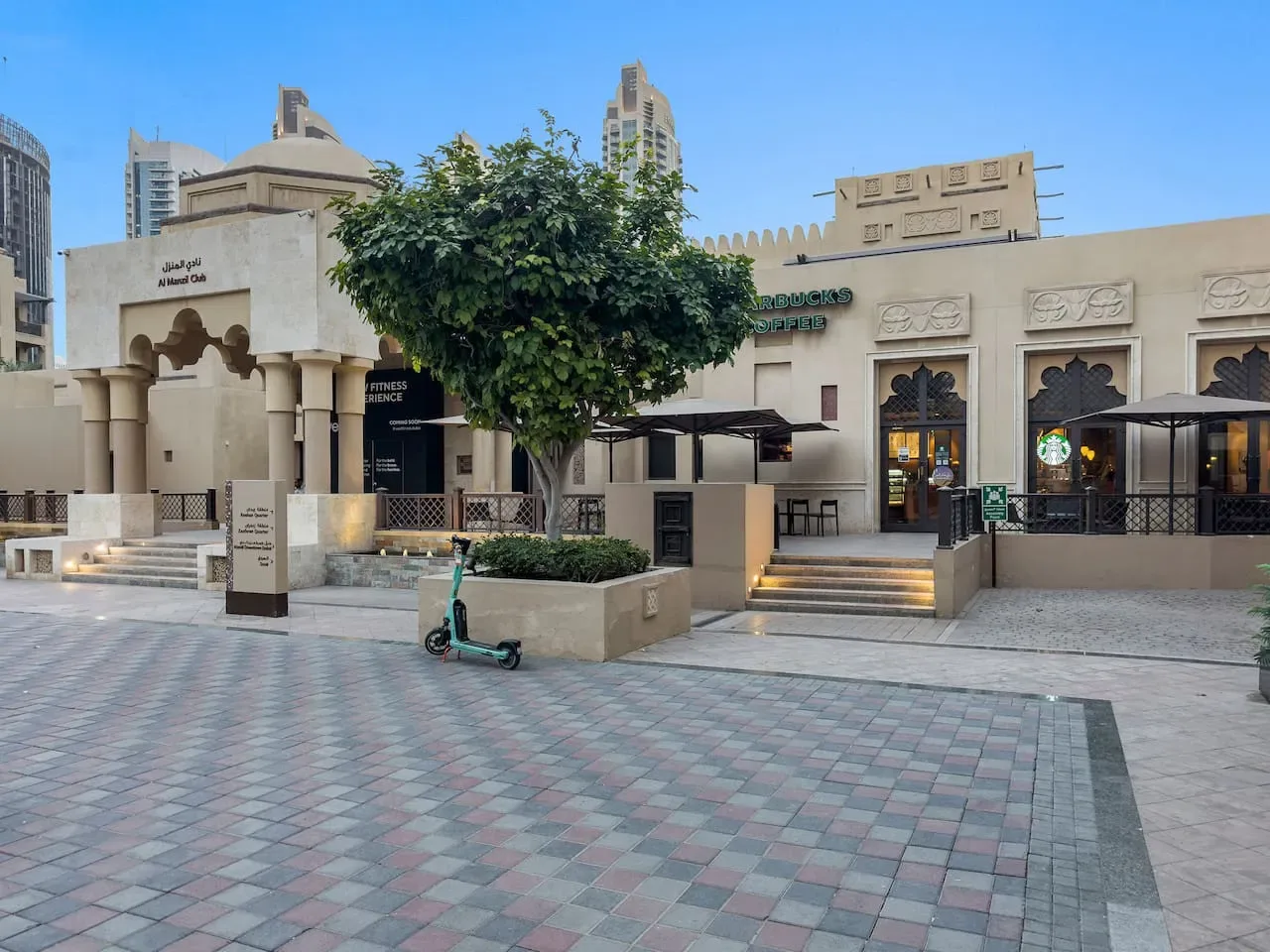 Una plaza peatonal pavimentada con un árbol frondoso y locales comerciales.