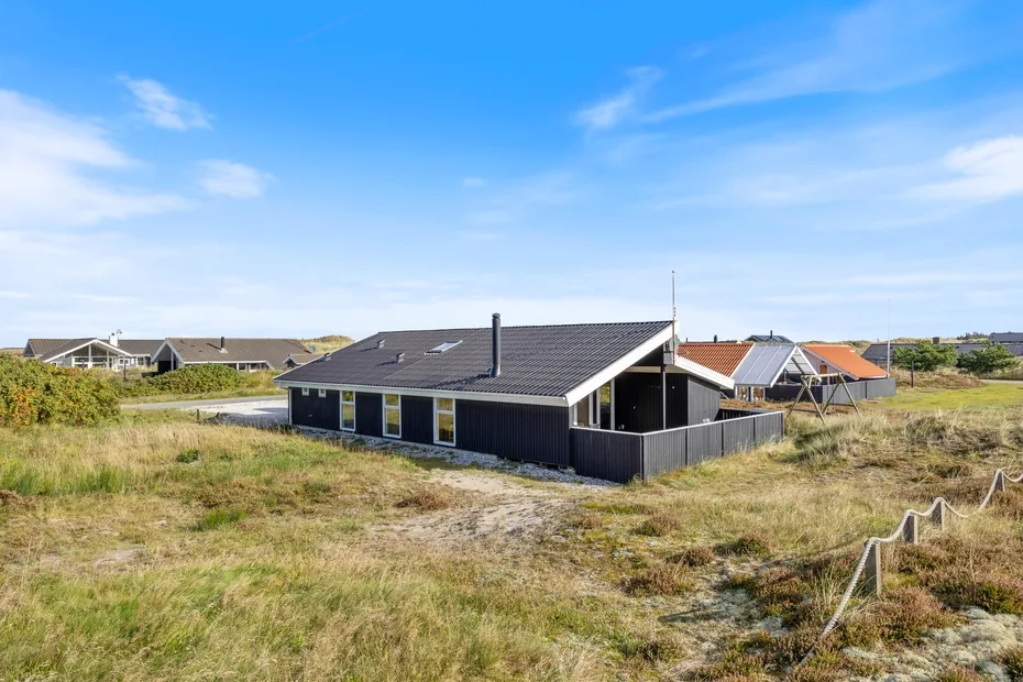 Una casa de techo oscuro se encuentra entre dunas cubiertas de hierba bajo un cielo azul.