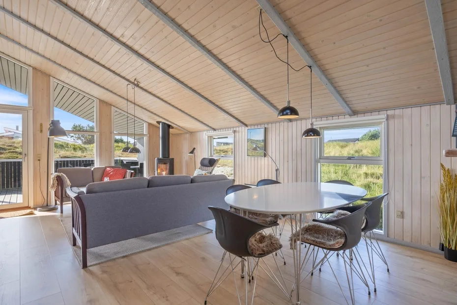 Una luminosa sala de estar y comedor diáfanos tiene grandes ventanas y techo de madera.