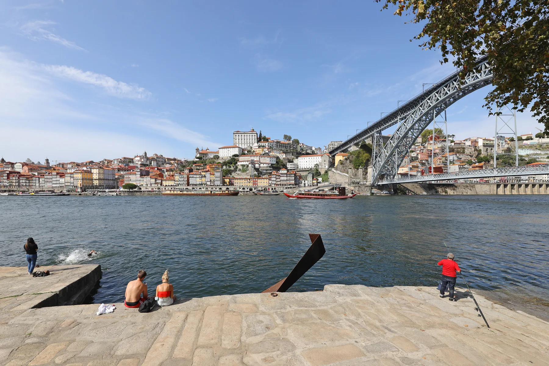 Riverside scene with people on a stone promenade beneath a large arched bridge.