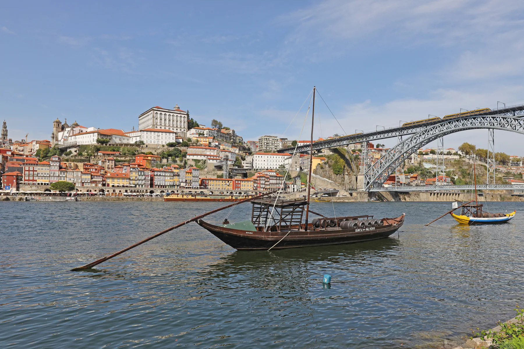 A wooden boat floats on the river, with a cityscape and arched bridge behind it.