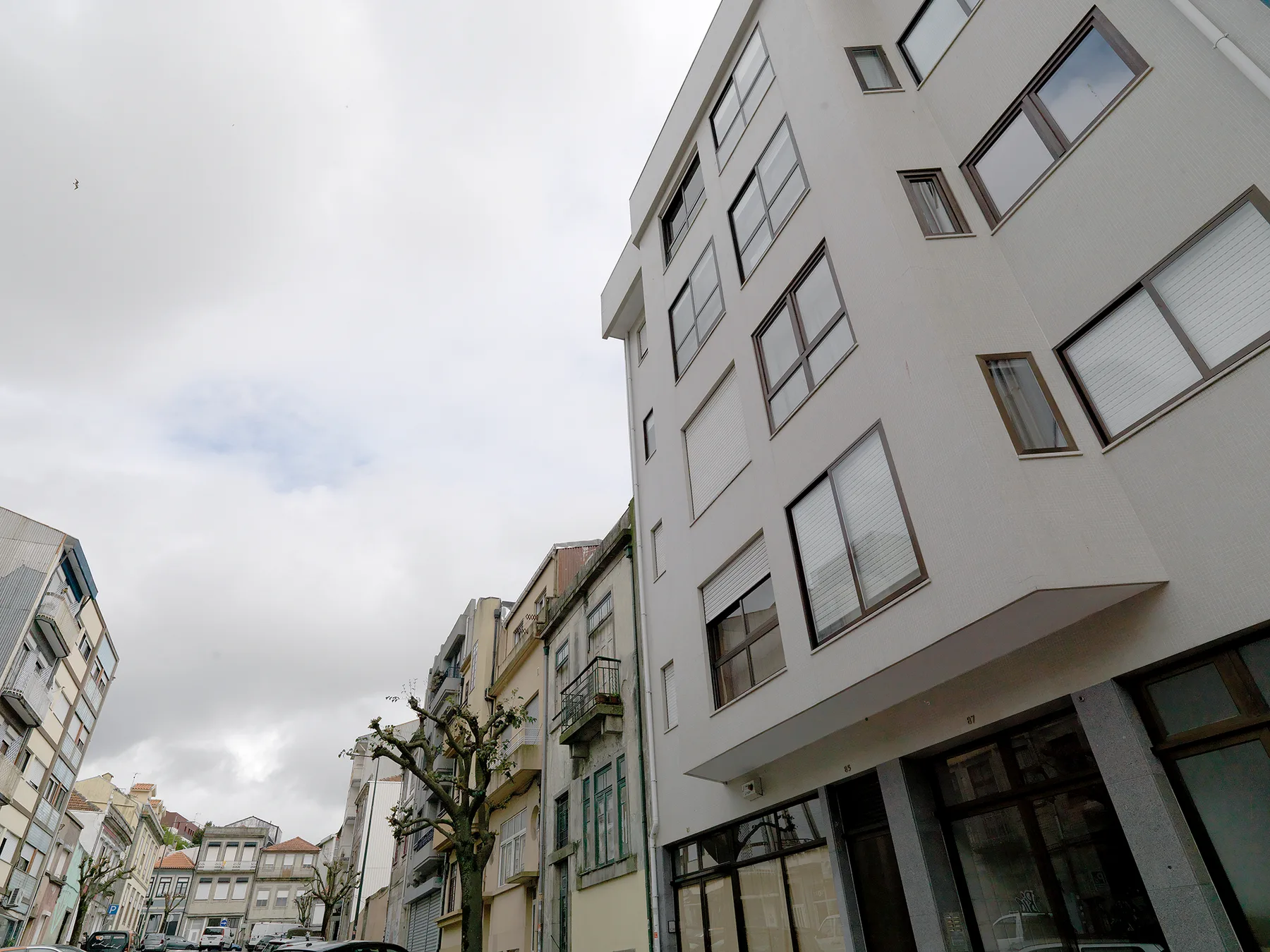 A street view shows a modern building beside older neighboring facades under a cloudy sky.