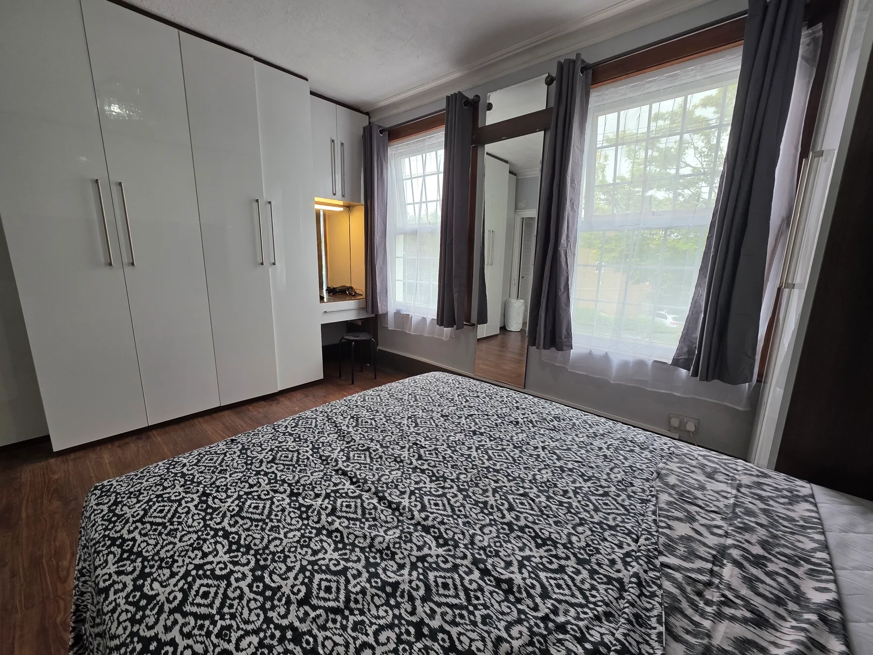 A bedroom with a patterned bedspread, white wardrobes, and tall curtained windows.
