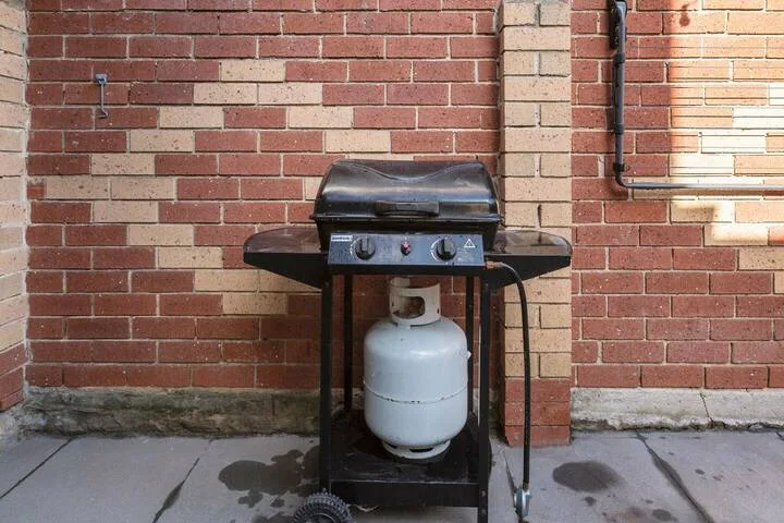 An outdoor gas grill stands against a brick wall on a paved patio.