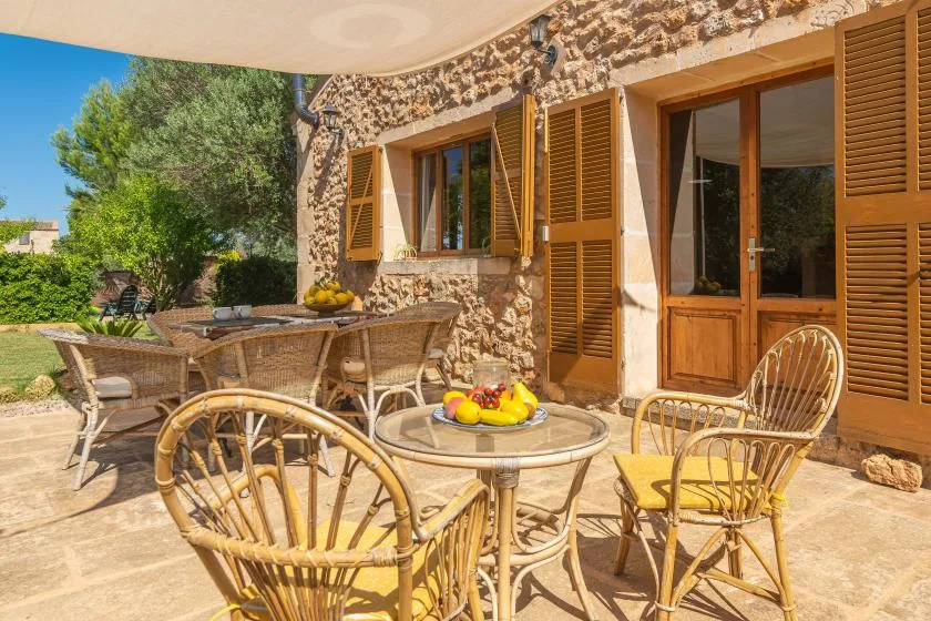 A sunny outdoor patio with wicker seating beside a stone wall and wooden shutters.