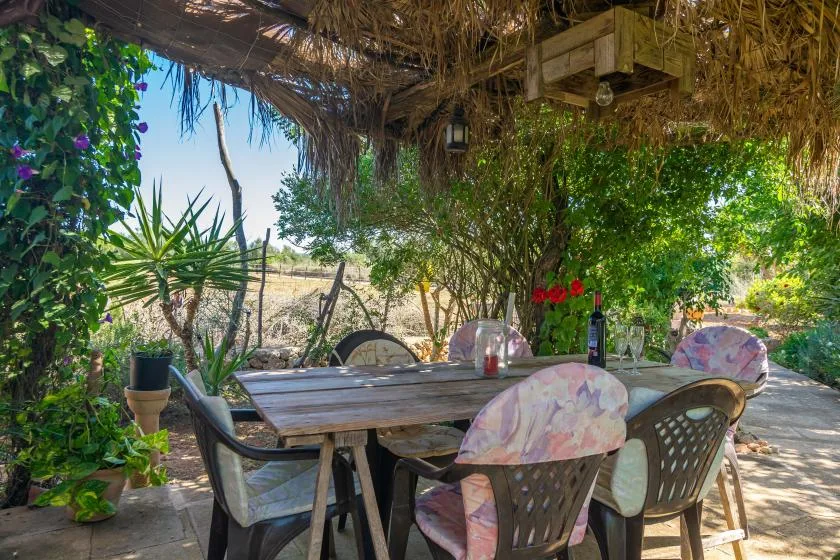 Una zona de comedor al aire libre está bajo un techo de paja, rodeada de vegetación.