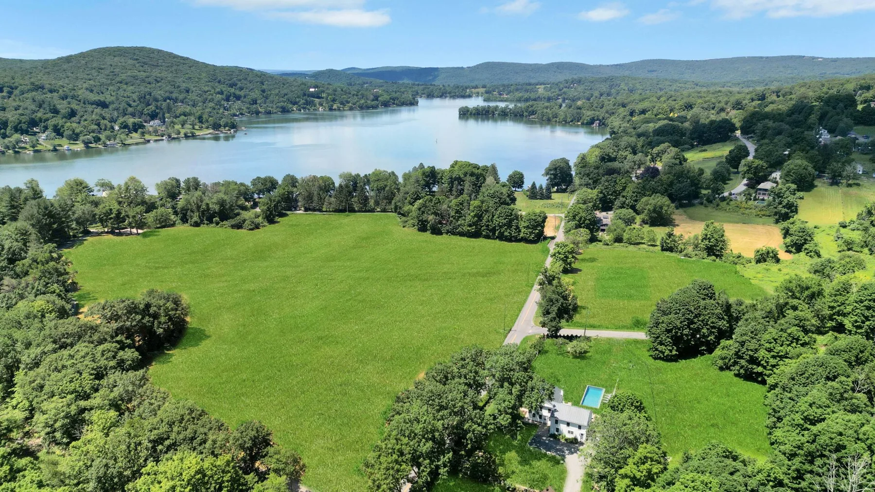 An aerial view of a broad green field beside a lake and wooded hills.