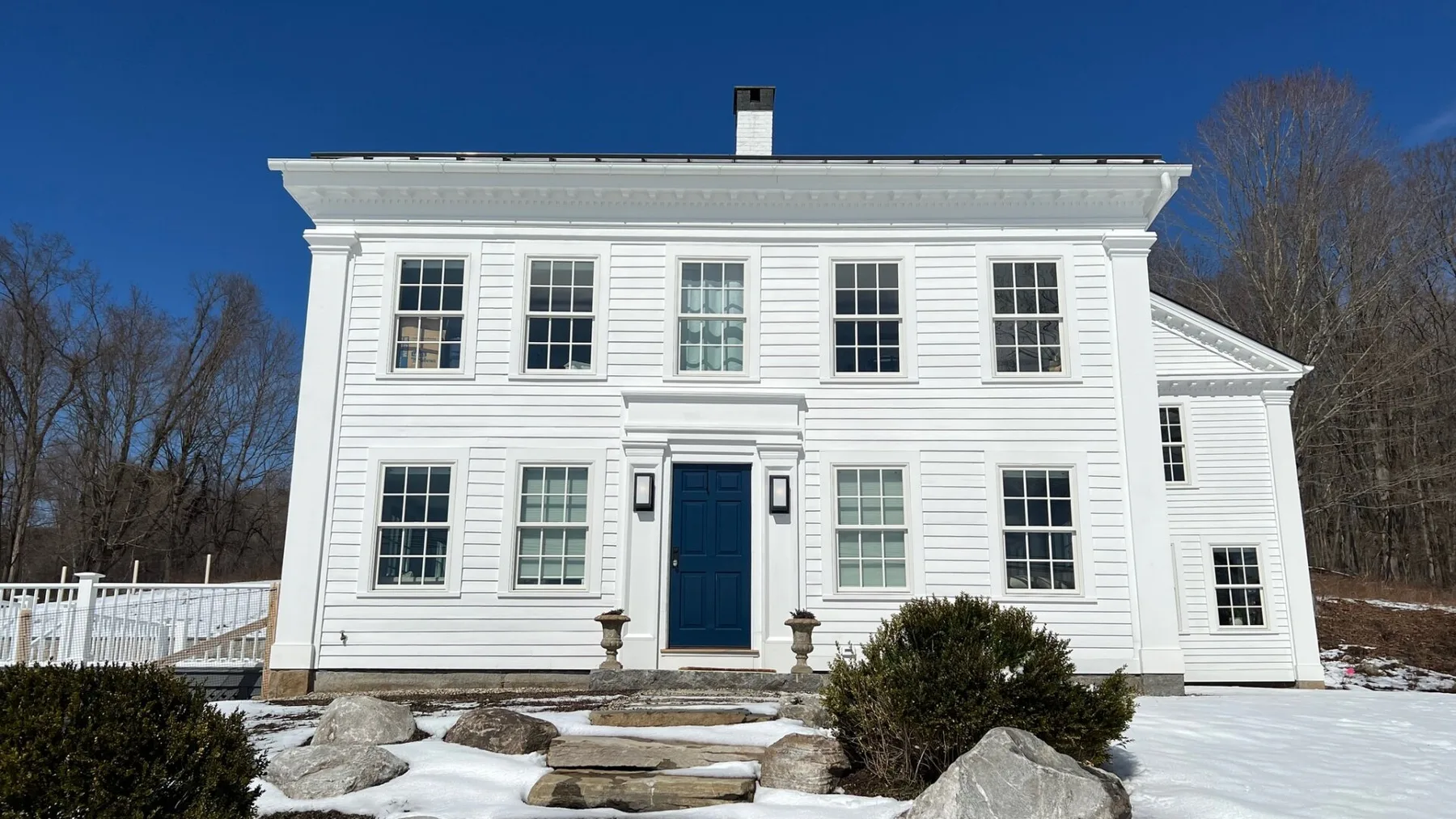 A white two-story house stands in a snowy yard beneath a clear blue sky.
