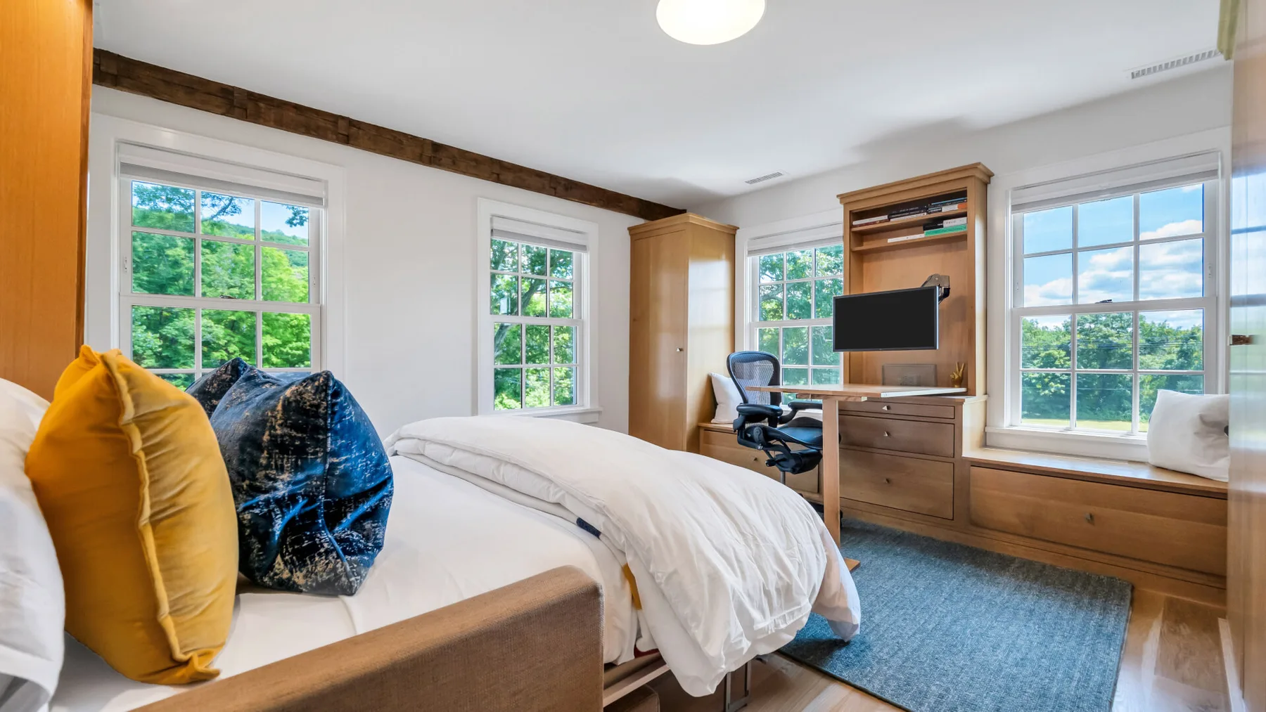 A bright bedroom with a bed, large windows, and a wooden desk.