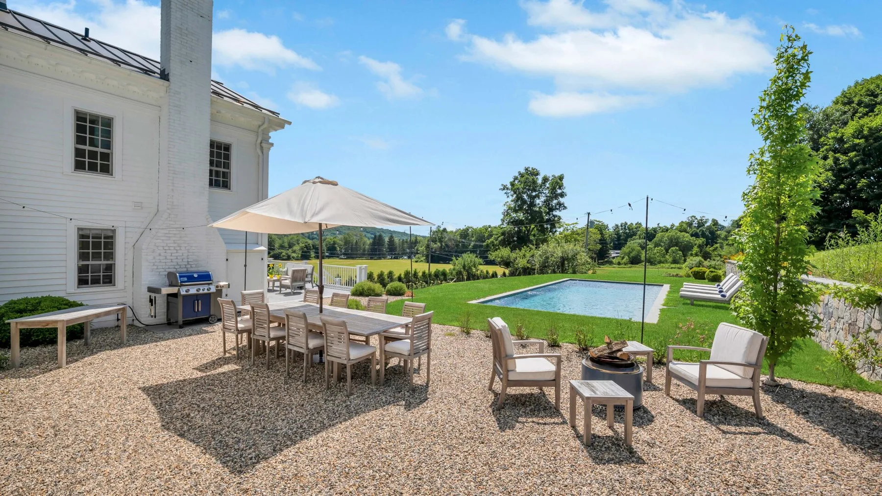 A gravel patio with outdoor seating and an umbrella overlooks a lawn and swimming pool.