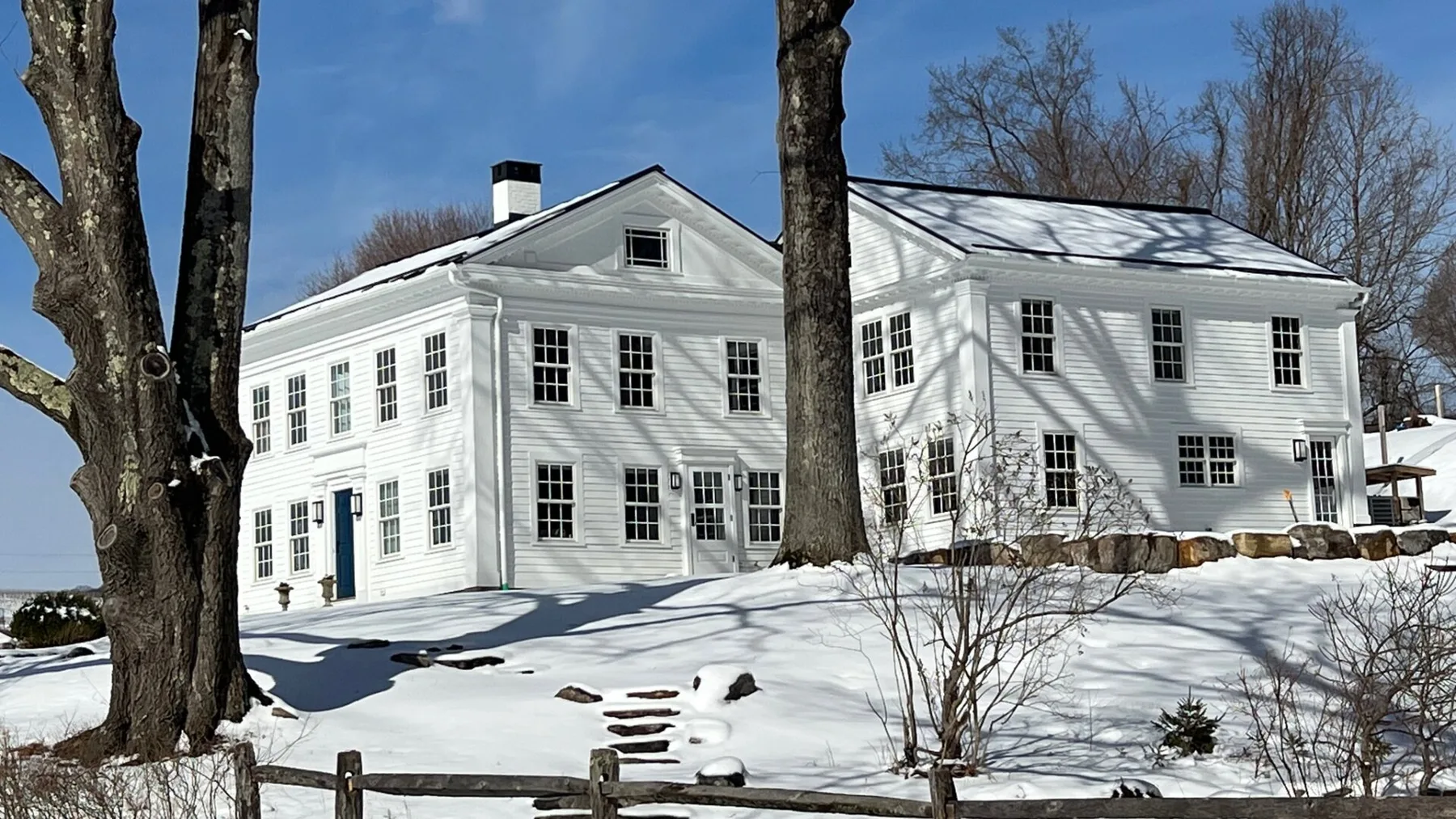 A large white house stands behind a snow-covered yard and bare trees.