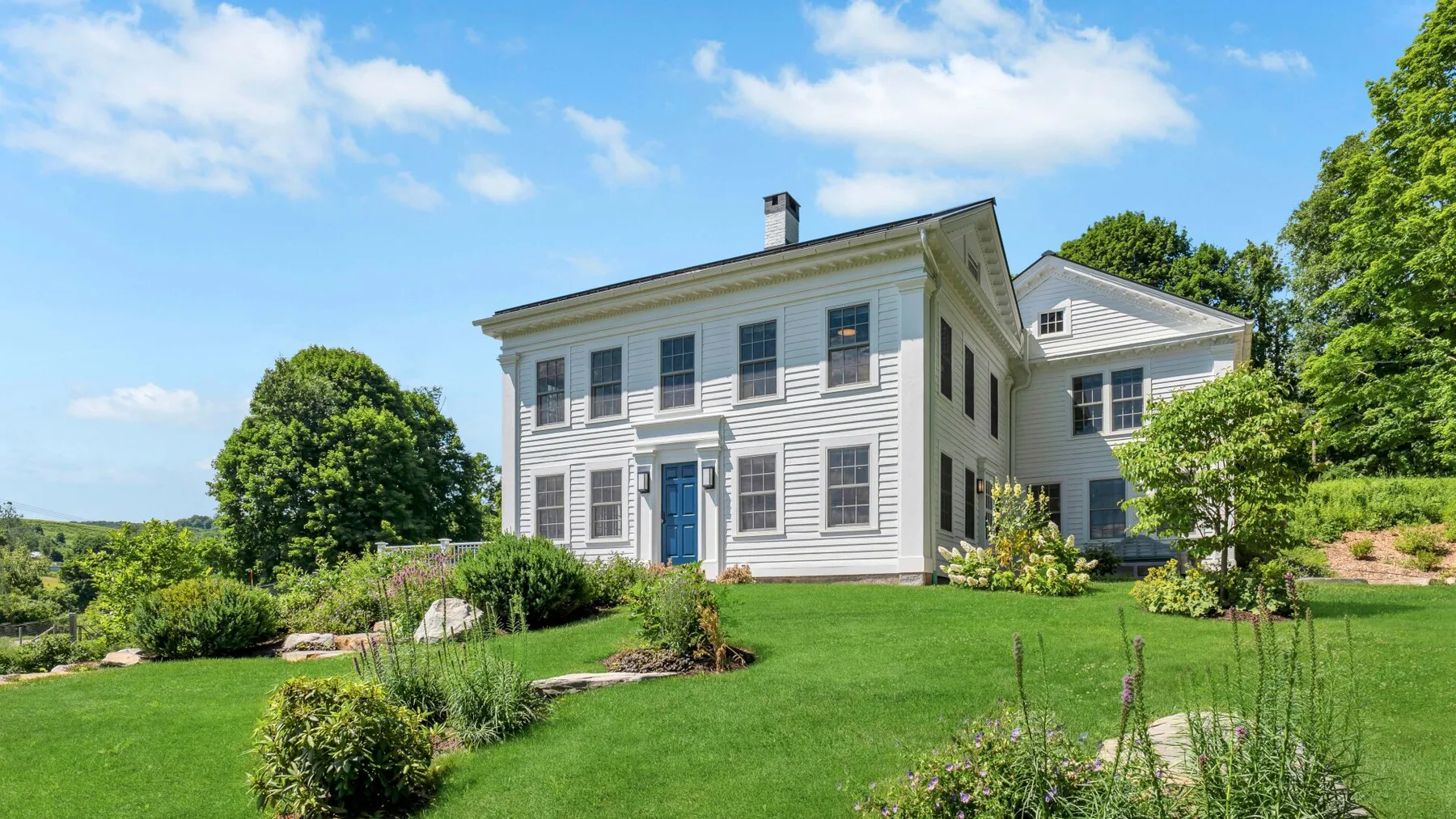 A white house stands amid a green lawn, shrubs, and trees.