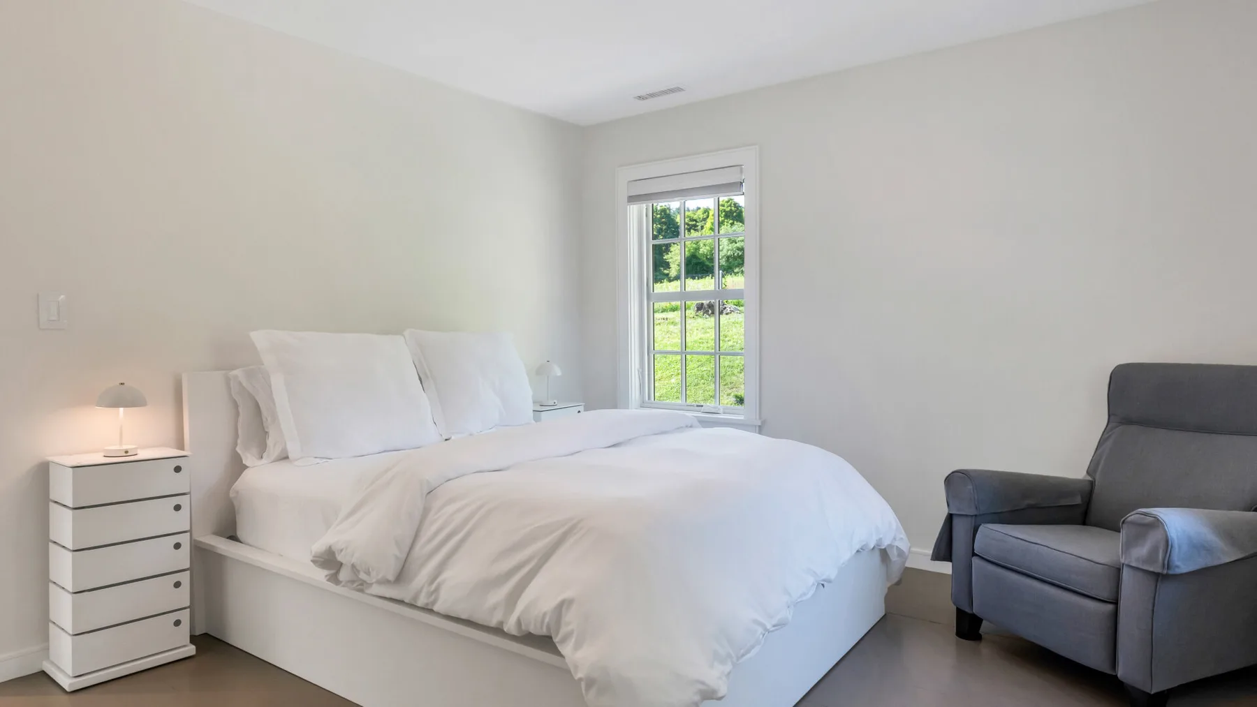 A bright bedroom with a neatly made bed and a gray armchair beside the window.