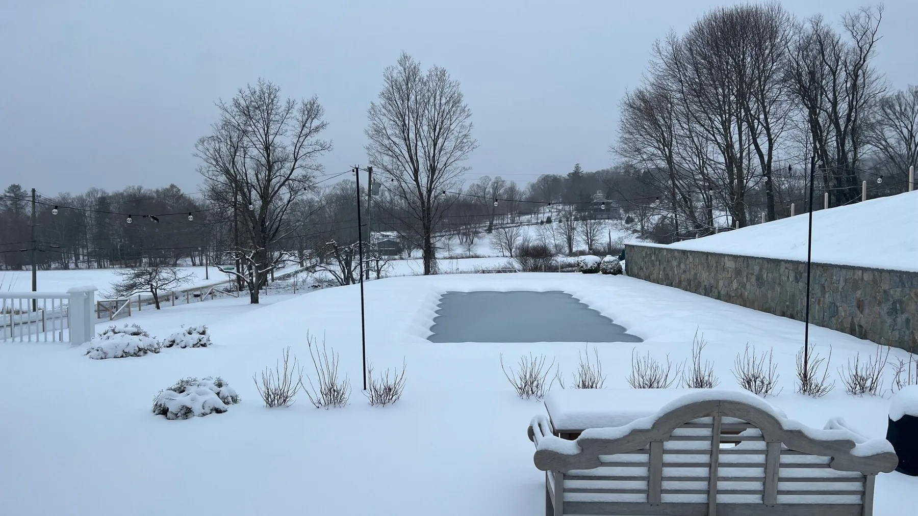 A snow-covered outdoor pool area with a stone wall and leafless trees.