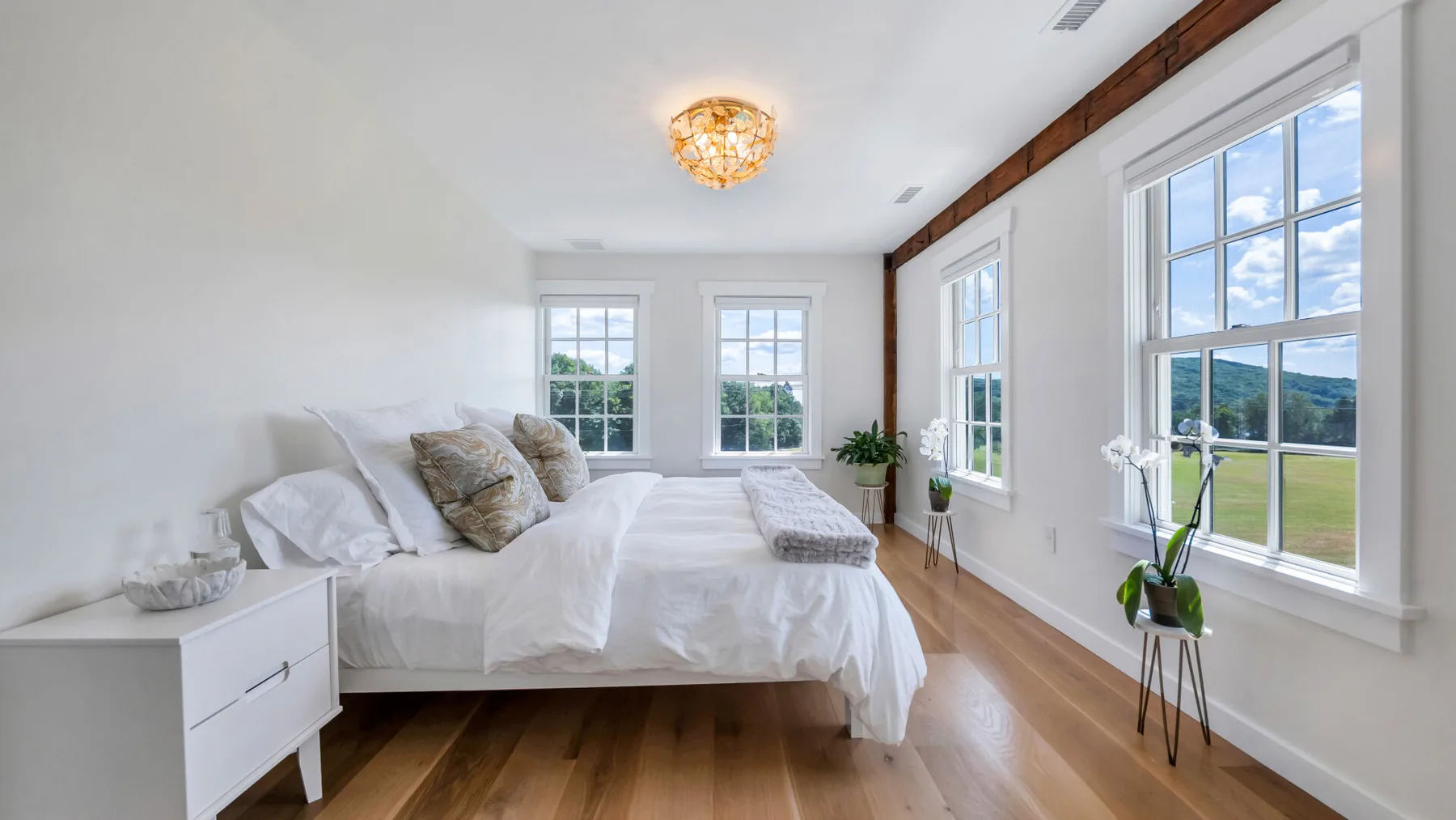 A bright bedroom has a bed, wood flooring, and large windows.