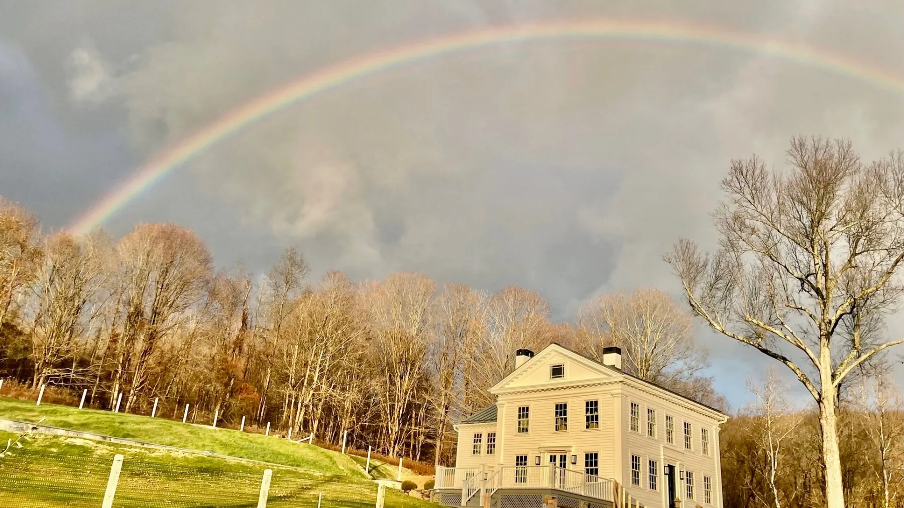 A large house stands beside a grassy slope beneath a rainbow.