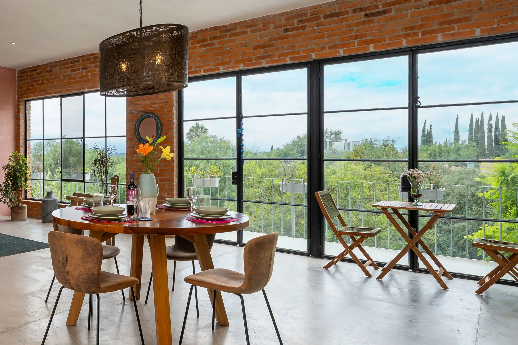 A dining area with a set table beside large windows overlooking greenery.