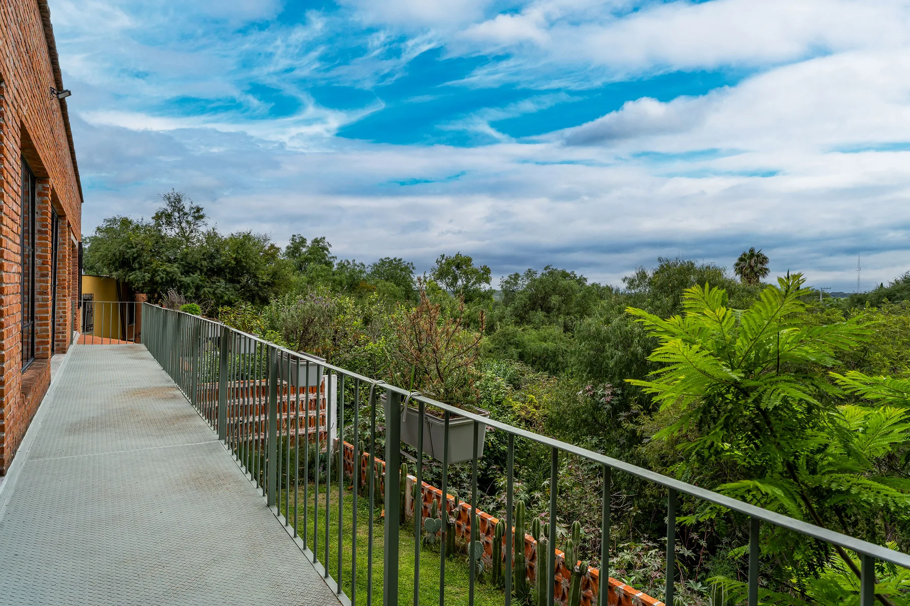 A balcony walkway overlooks leafy greenery beneath a partly cloudy sky.