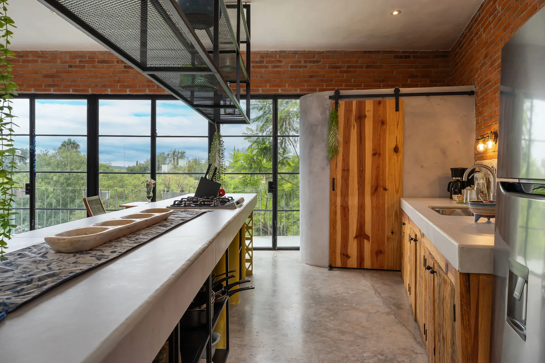 A kitchen with a long island, wooden cabinetry, and large windows overlooking greenery.