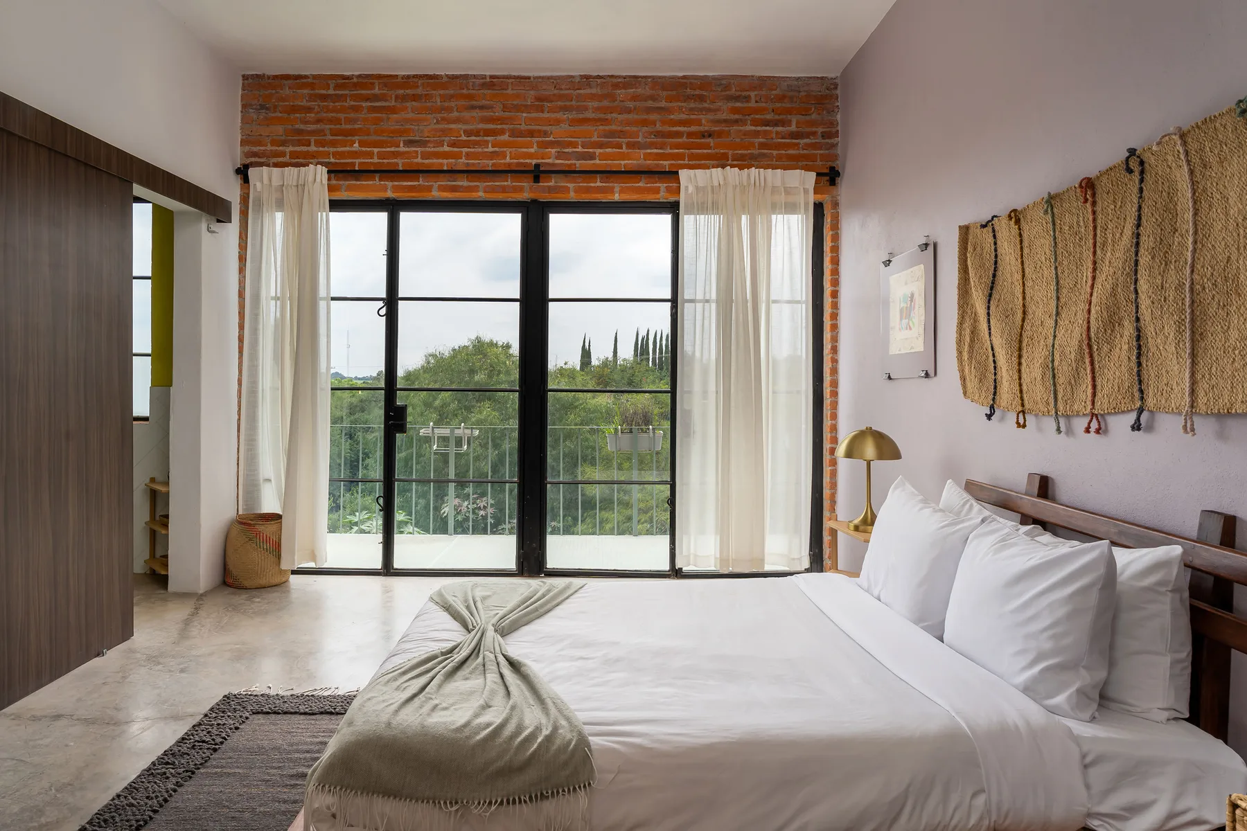 A bedroom with a bed beside tall windows overlooking greenery.