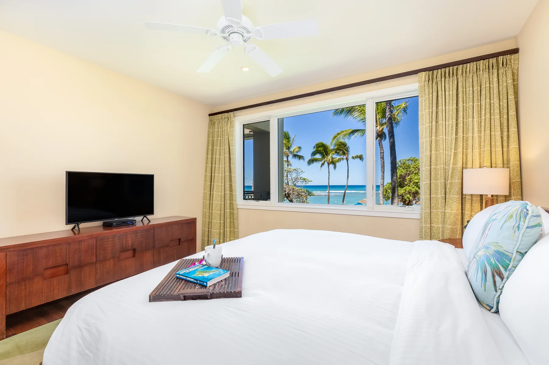 Un dormitorio con una cama grande frente a una ventana con vista a palmeras y al mar.