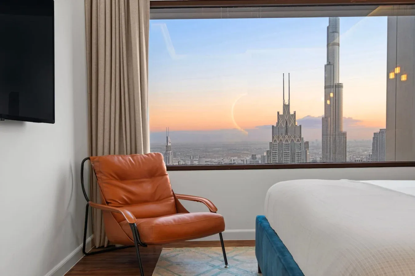 A bedroom with a large window overlooking city buildings at sunset.