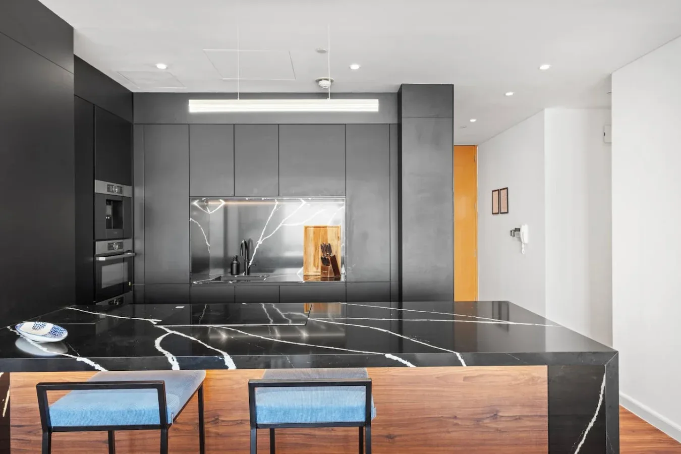 A modern kitchen with dark cabinetry, a large island, and wood-front seating.
