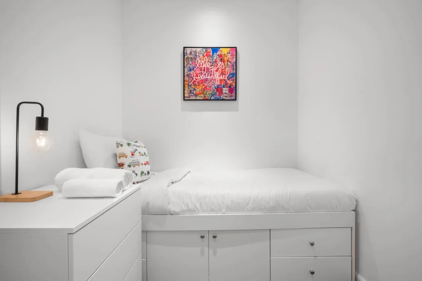 A compact bedroom with a white bed, built-in storage, and colorful wall art.