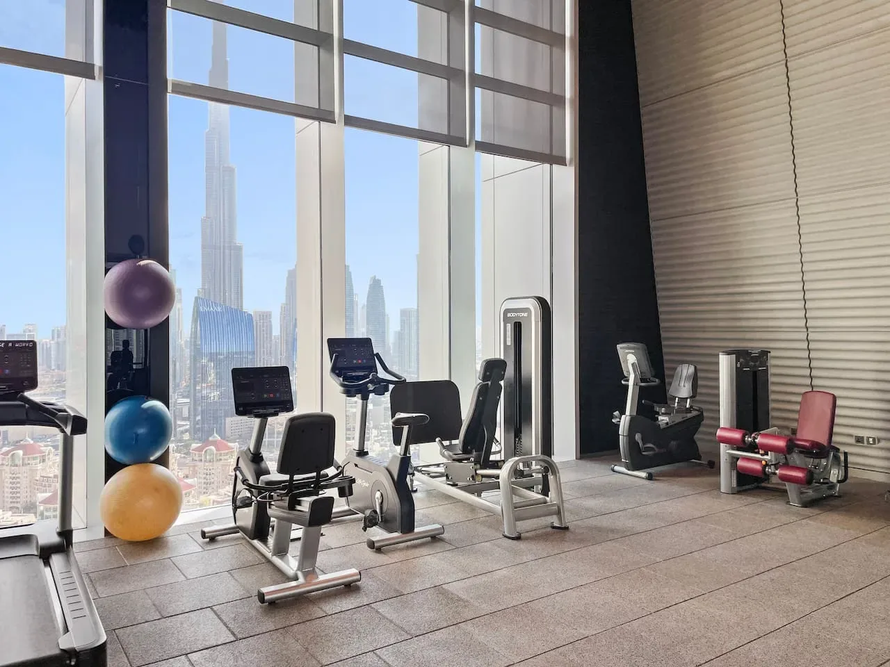 A gym with exercise machines beside floor-to-ceiling windows overlooking a city skyline.
