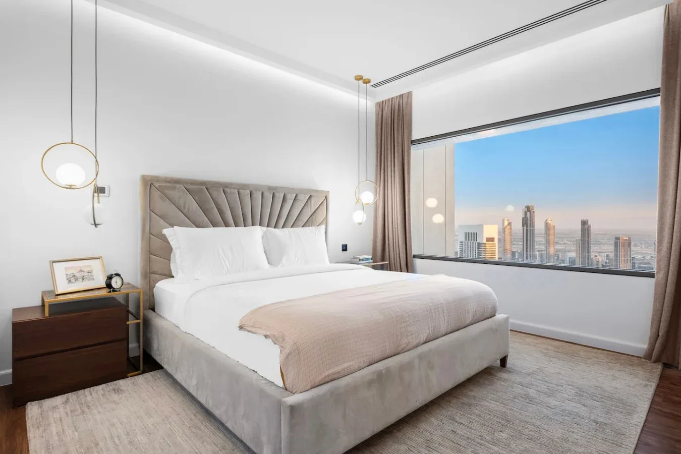 A bright bedroom has a neatly made bed beside a large window overlooking the city.