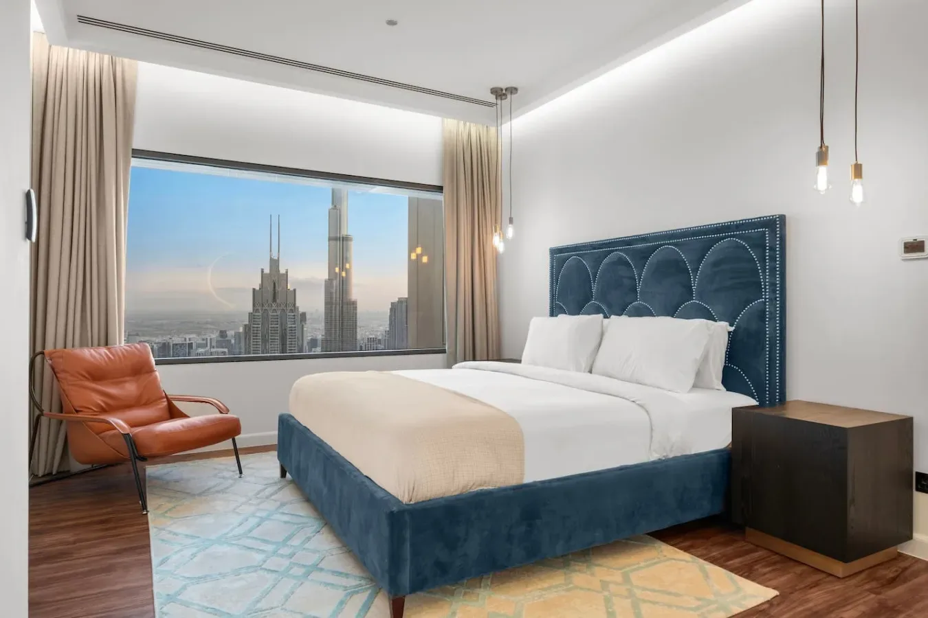 A bedroom with a bed, an orange lounge chair, and a city view through the window.