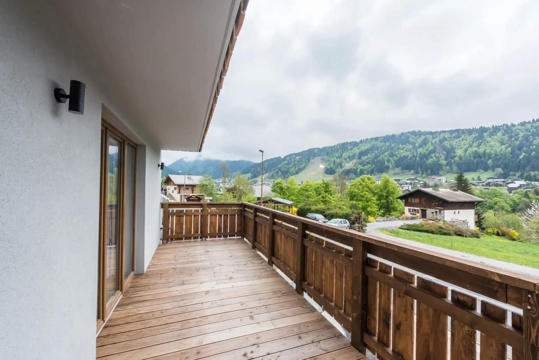 A wooden balcony with mountain views overlooks a green valley and nearby buildings.