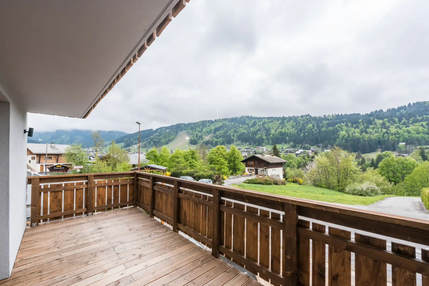 A wooden balcony overlooks green hills and scattered buildings beneath a cloudy sky.
