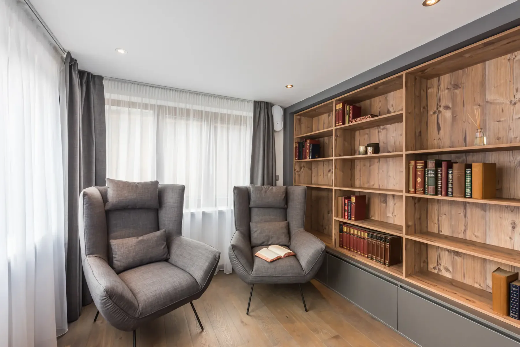 A living area with two gray armchairs beside a wooden bookshelf.
