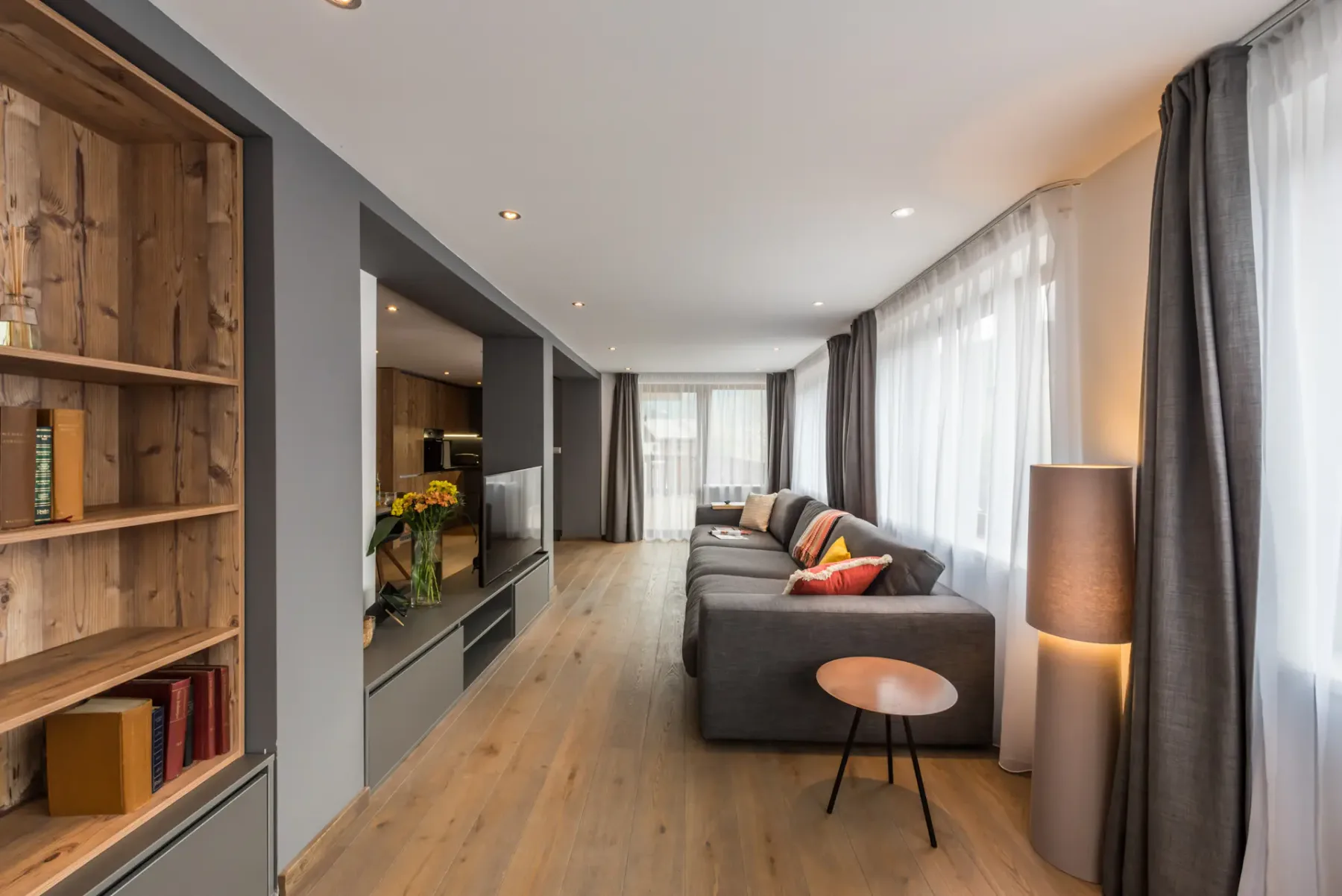 A living room with a sectional seating area beside large curtained windows.