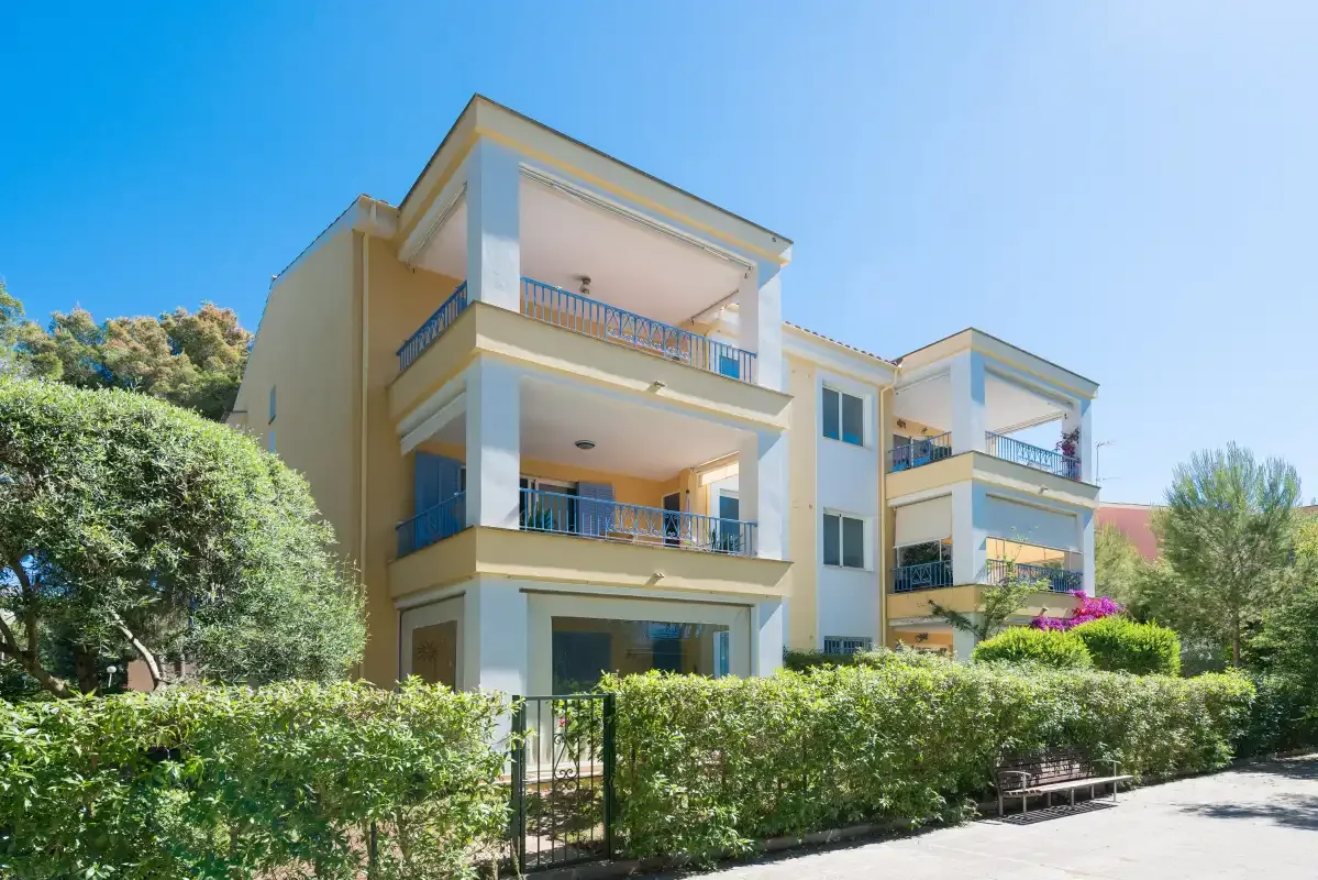 A light-colored apartment building with balconies stands behind a leafy hedge.
