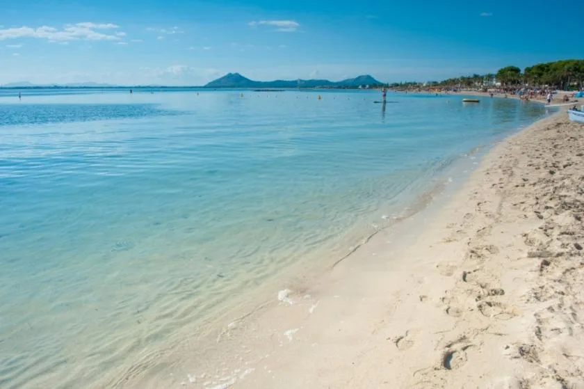 A sandy beach curves along clear, shallow blue water beneath a bright sky.