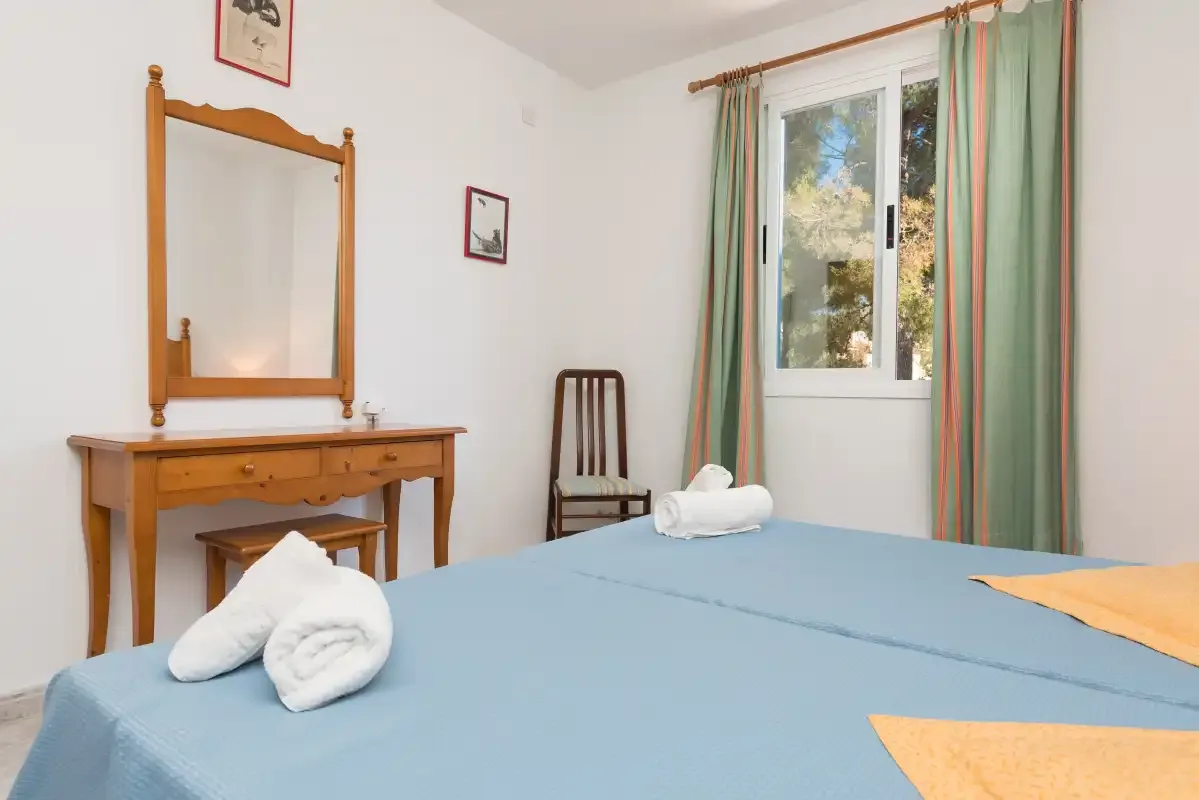 A bedroom with a blue bedspread, wooden dressing table, and curtained window.