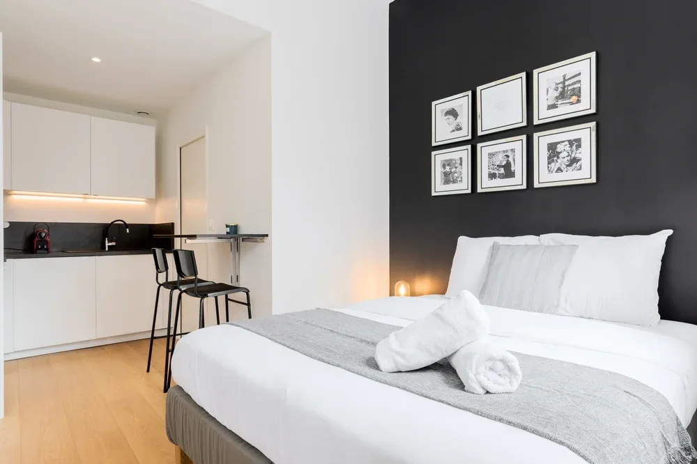 Dormitorio con ropa de cama blanca y una pequeña zona de cocina al fondo.