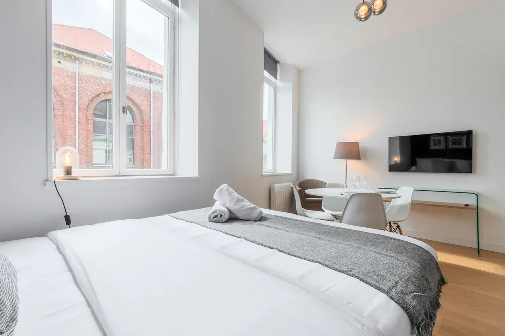 A bright bedroom with a bed, a small dining area, and a wall-mounted television.