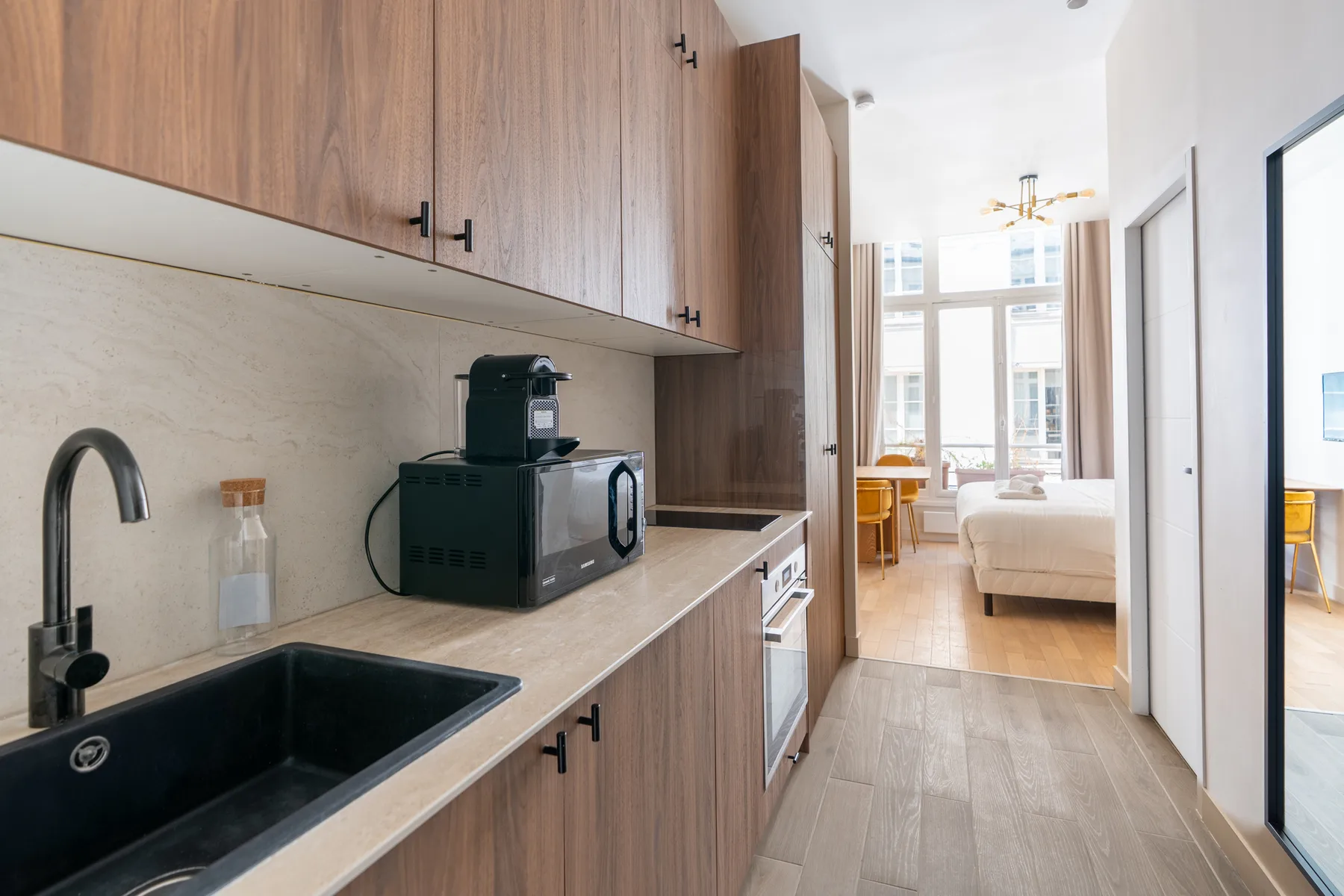 A kitchenette with a sink and microwave opens toward a bright bedroom.
