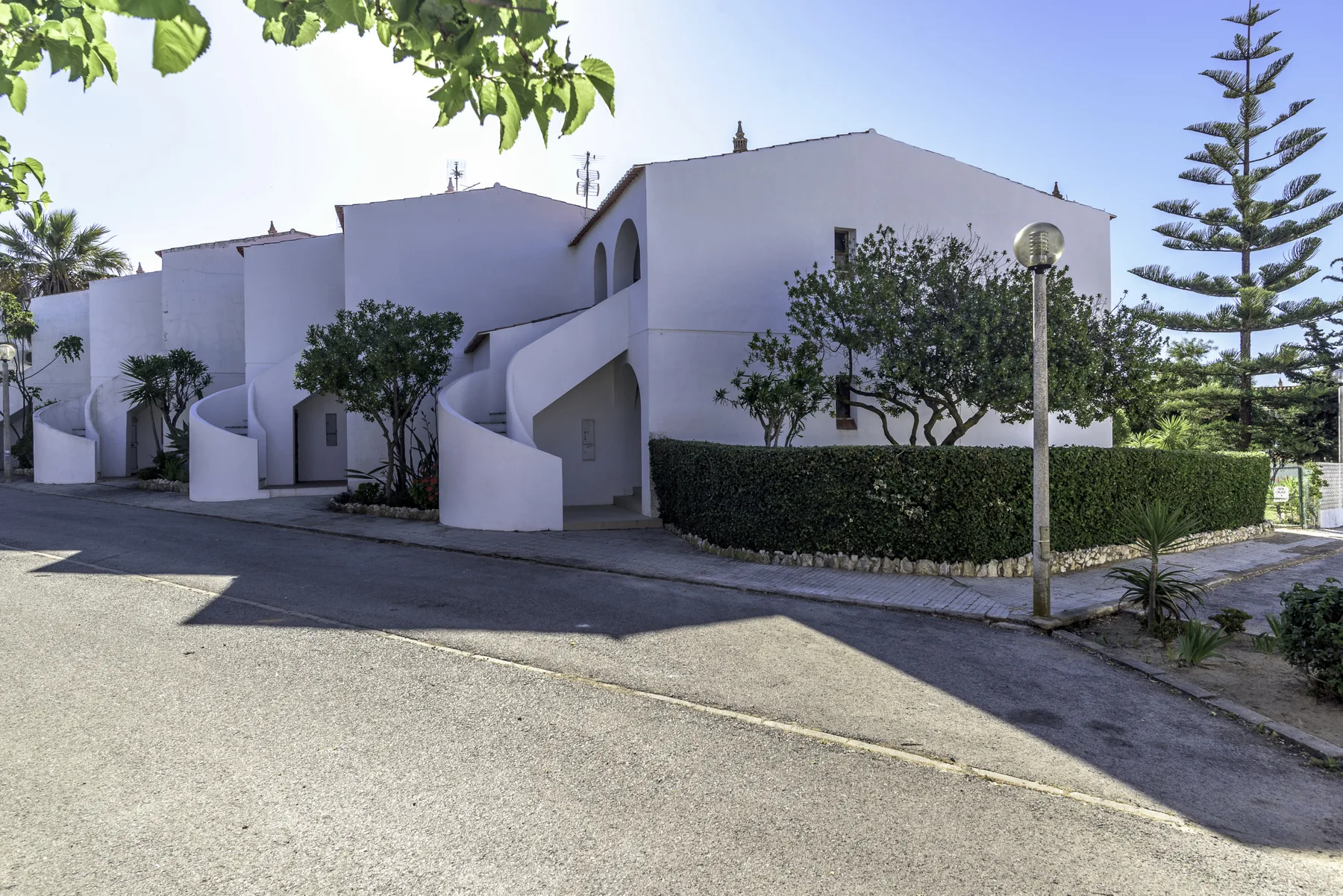 Edificio blanco con escaleras exteriores y vegetación junto a una calle pavimentada.