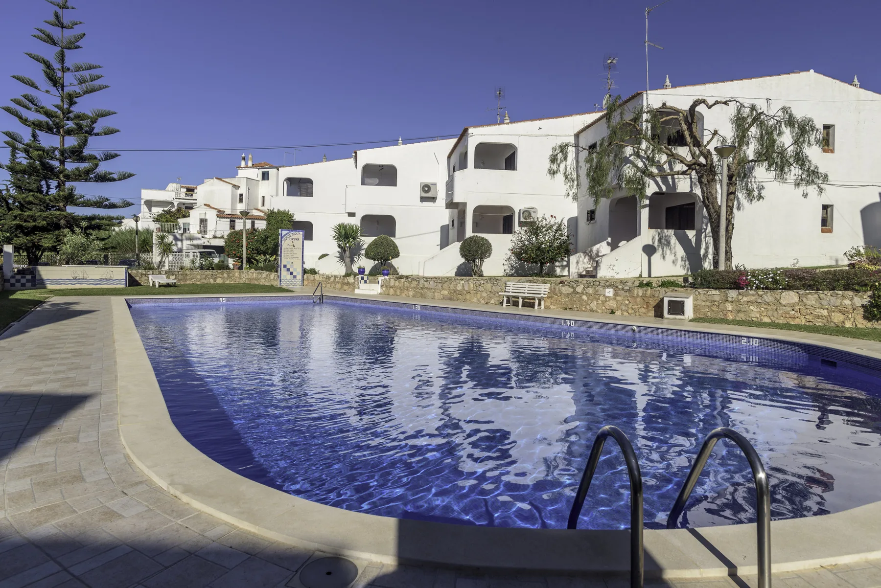 Una piscina está frente a edificios blancos y un sendero ajardinado.