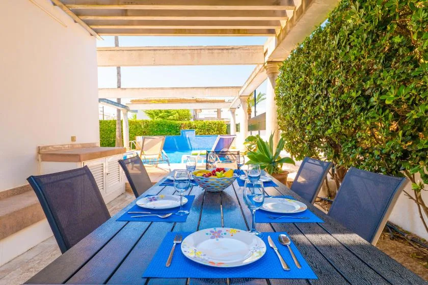 Comedor al aire libre con una mesa puesta bajo una pérgola, junto a una piscina.