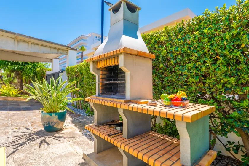 Una barbacoa de ladrillo al aire libre está junto a un patio pavimentado y un seto frondoso.