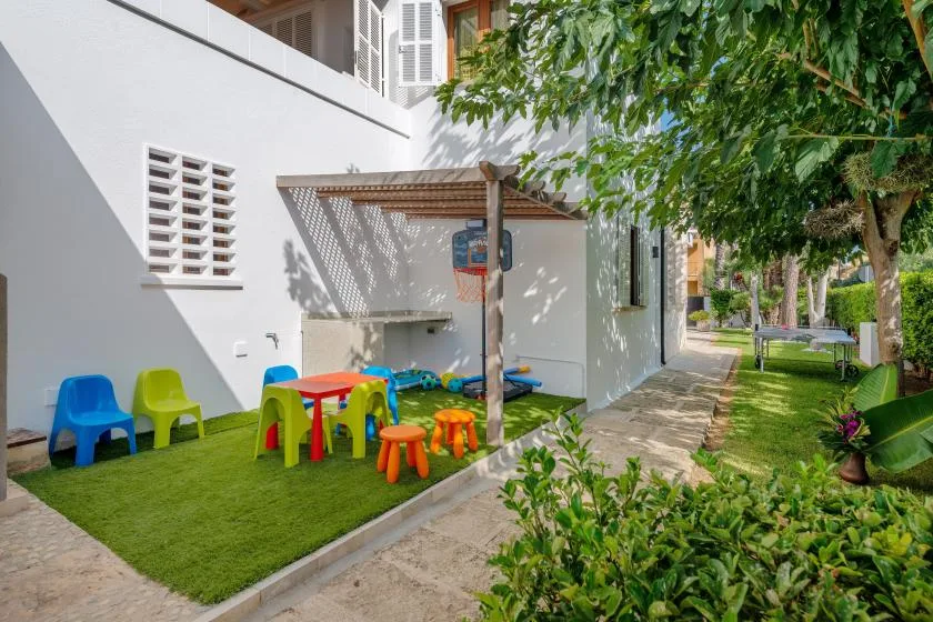 Una zona de juegos al aire libre junto a un edificio blanco, con sillas pequeñas y un sendero sombreado.